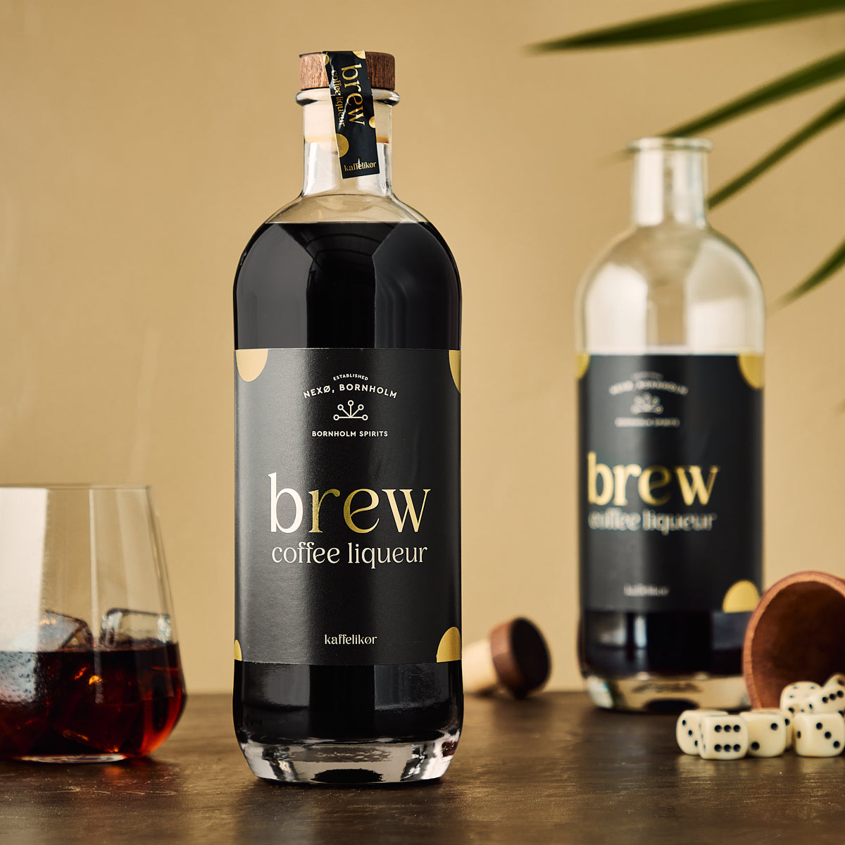 Flasche Brew Kaffeelikör im Vordergrund, zweite Flasche im Hintergrund, Glas mit Likör und Würfel auf Tisch.
