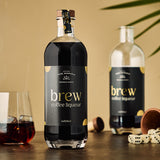 Flasche Brew Kaffeelikör im Vordergrund, zweite Flasche im Hintergrund, Glas mit Likör und Würfel auf Tisch.