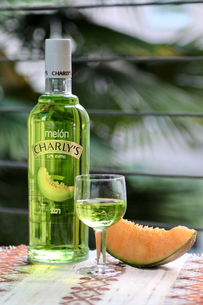 Flasche Charly's Melón alkoholfrei, mit Glas grünem Getränk und Melonenscheibe, vor unscharfem Hintergrund.