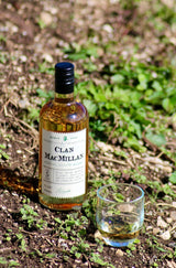 Eine Flasche Clan MacMillan Blended Scotch Whisky steht auf einem naturbelassenen Boden neben einem Glas Whisky. Im Hintergrund sind grüne Pflanzen zu sehen.