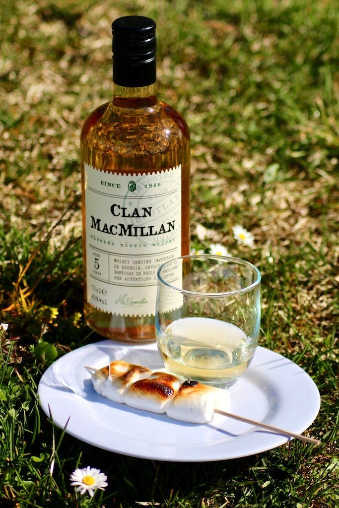 Eine Flasche Clan MacMillan Blended Scotch Whisky steht auf einer Wiese neben einem Glas Whisky und einem Teller mit gerösteten Marshmallows auf einem Holzspieß.