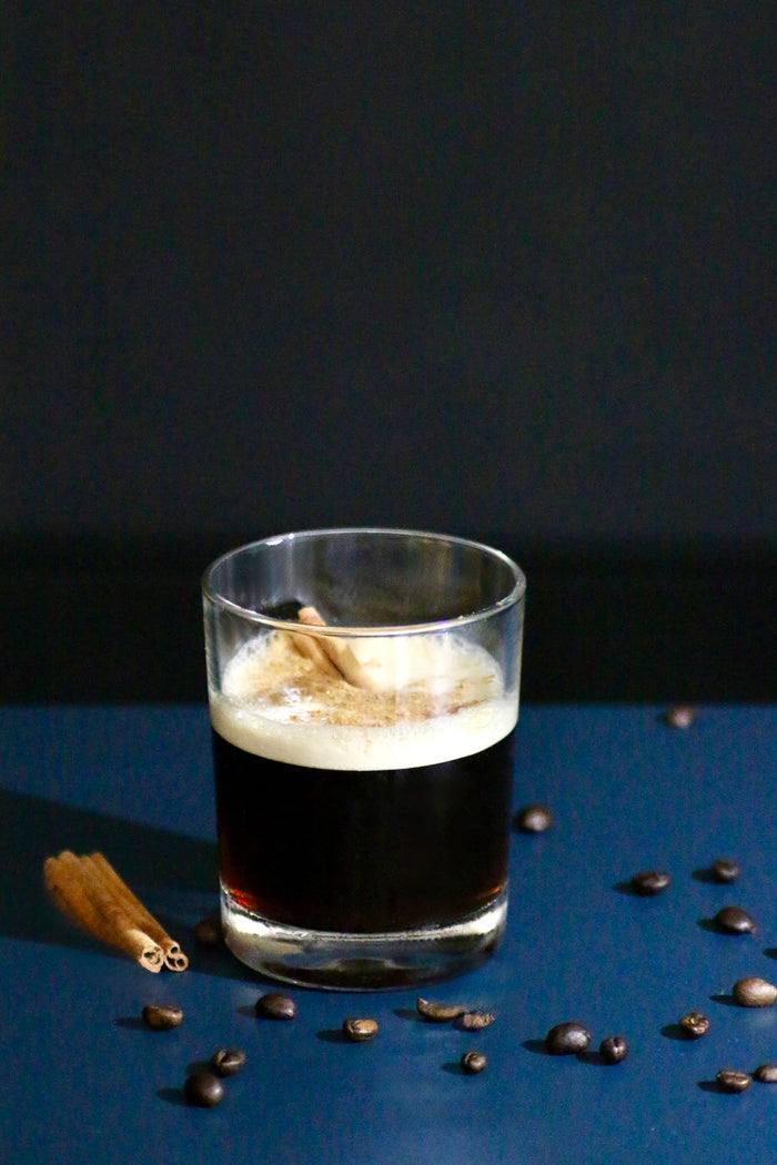 Coffee Shadow Cocktail im Rocks-Glas mit Zimtrinde, umgeben von Kaffeebohnen auf dunkler Oberfläche.