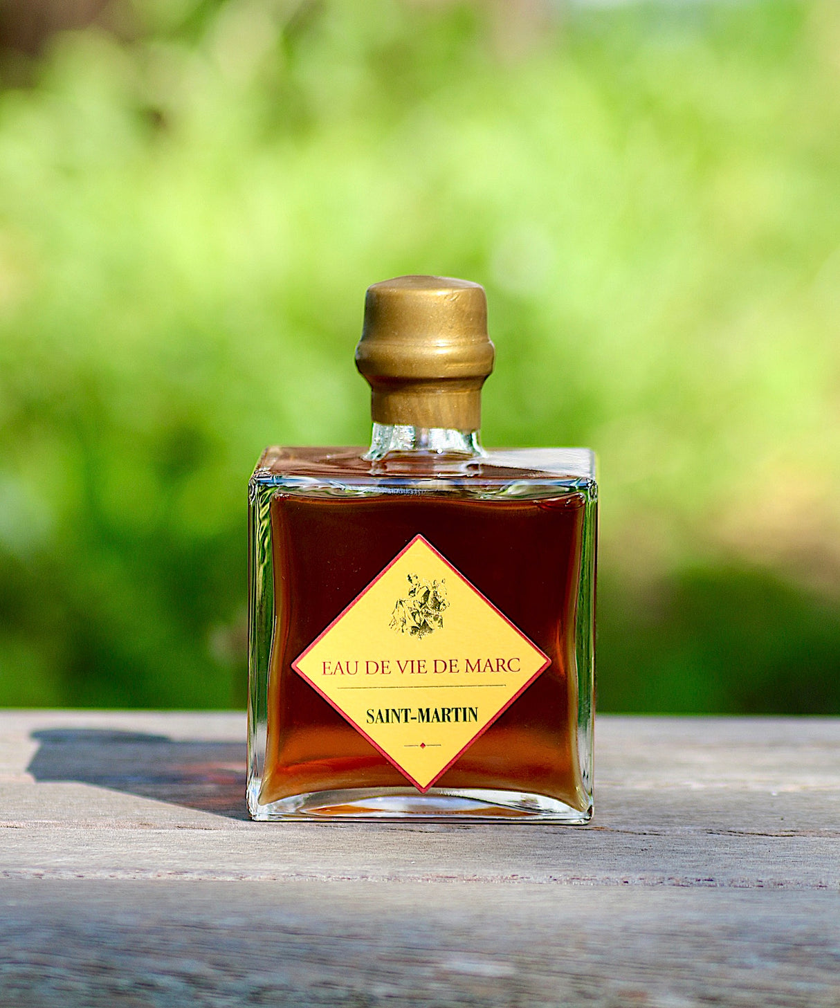 Flasche Eau de Vie de Marc Saint-Martin mit goldfarbenem Etikett auf einem Holztisch, im Hintergrund eine verschwommene grüne Landschaft.