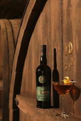 Flasche Grégorienne Kräuterlikör vor großen Holzfässern, daneben ein Bijou-Cocktail im Glas mit Zitronenzeste.