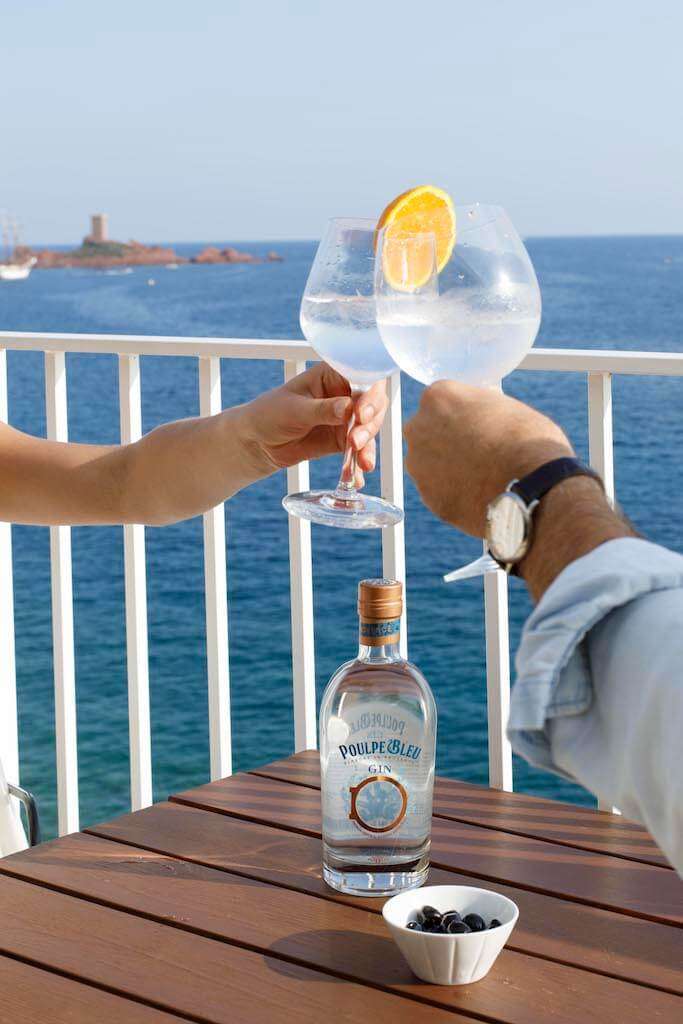 Zwei Personen stoßen mit Gin Tonic Gläsern an, im Hintergrund Meer und Felsen, auf dem Tisch Poulpe Bleu Gin Flasche und Oliven.
