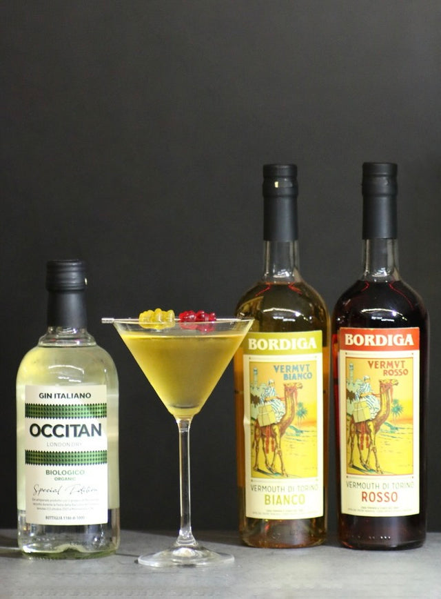 Flaschen von Occitan Gin und Bordiga Vermouth neben einem goldgelben Golden Ermine Cocktail im Martiniglas mit Gummibärchen-Garnitur.