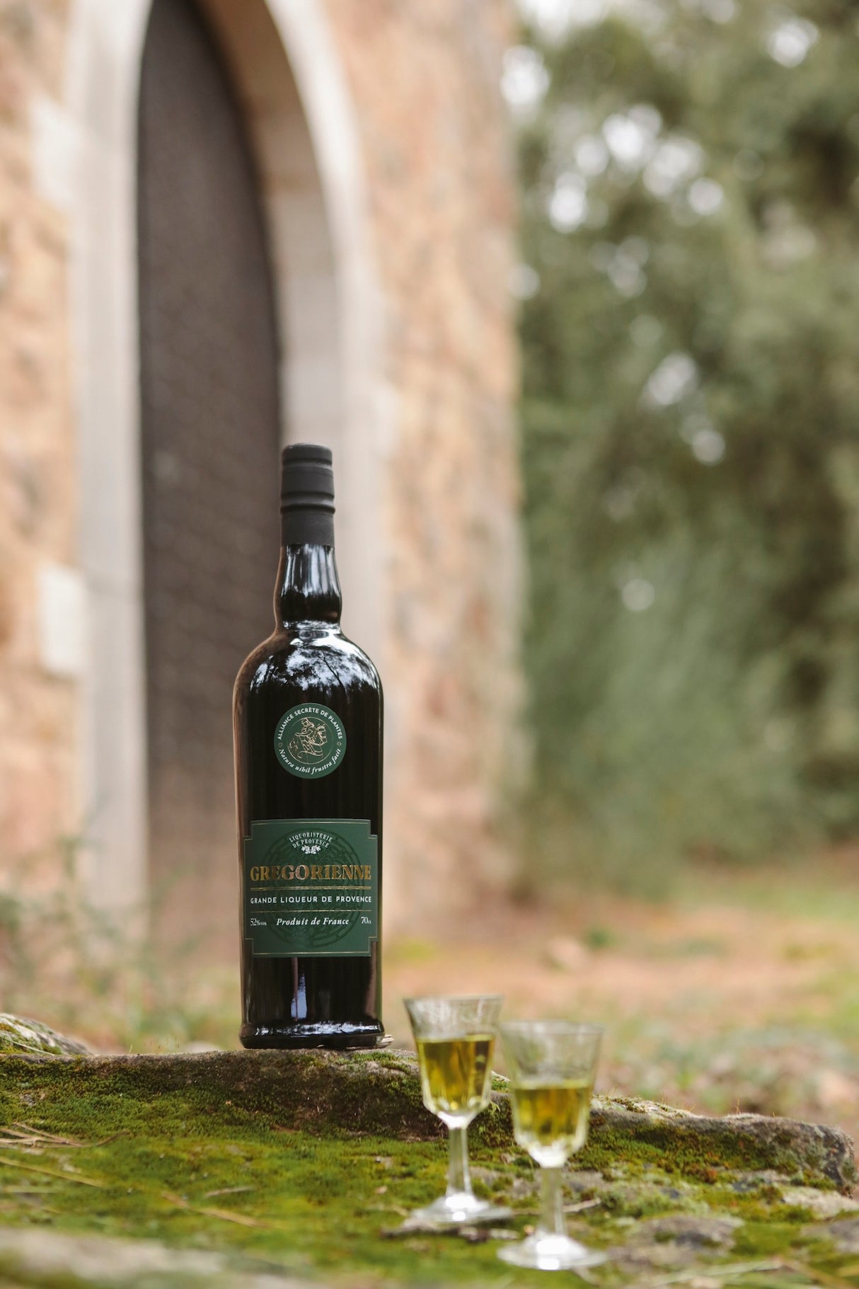 Gregorienne Kräuterlikör aus der Provence mit zwei gefüllten Gläsern vor Steinbogen und Gartenkulisse
