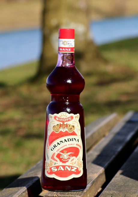 Flasche Sanz Grenadine Sirup 1L auf einer Holzbank, mit einem unscharfen Baum und einem Fluss im Hintergrund.