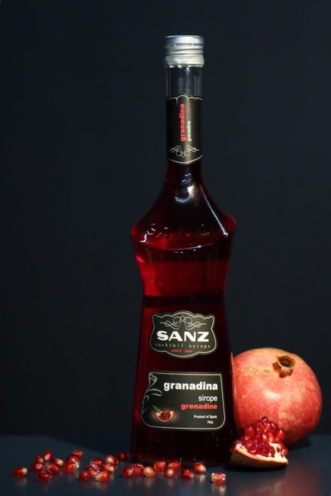 Flasche Sanz Grenadinesirup, umgeben von Granatapfelkernen und einem halben Granatapfel