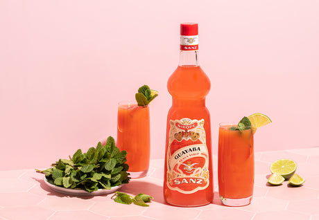 Flasche Guaven-Sirup von Sanz mit zwei Gläsern Guave-Limette-Spritz, frischer Minze und Limettenstücken vor rosa Hintergrund.