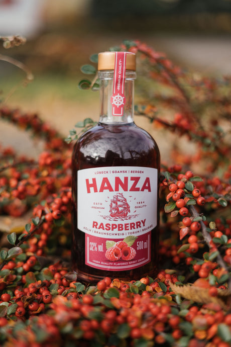 Hanza Raspberry Spirit Drink Flasche umgeben von roten Beeren und Herbstlaub. Rotes Etikett mit Segelschiff-Motiv.