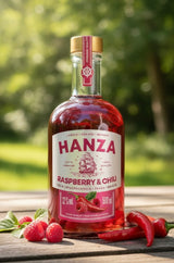 Hanza Himbeere & Chili Flasche auf Holztisch in sonniger Natur, dekoriert mit frischen Himbeeren und roten Chilischoten.