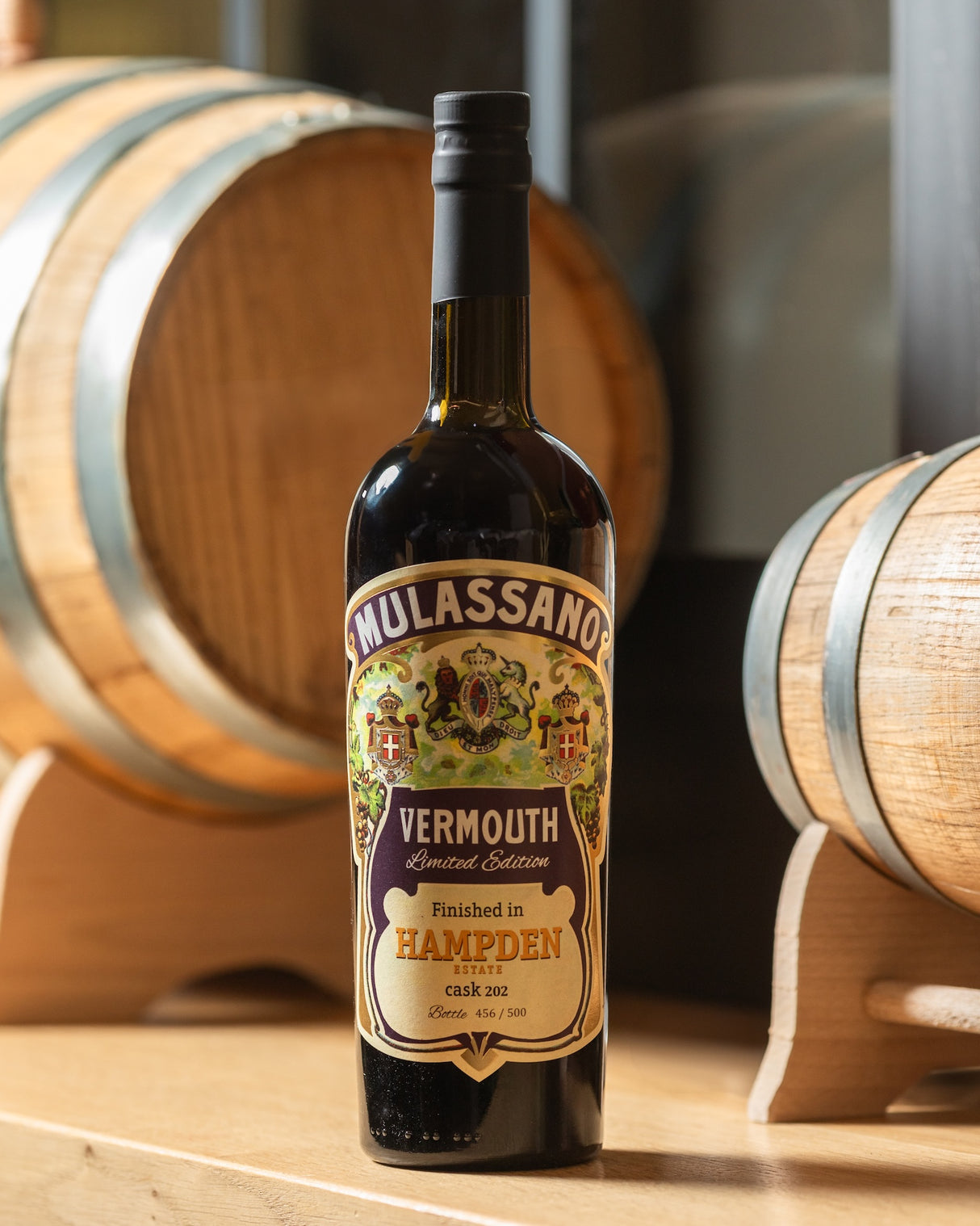 Mulassano Vermouth Flasche auf Holzregal vor Eichenfässern. Etikett: Finished in Hampden Estate cask 202, Bottle 456/500.