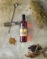 Flasche Bordiga Bitter Rosso, umgeben von Cocktailwerkzeugen und Botanicals wie Wacholderbeeren, Wurzeln und Samen auf einer Fläche.