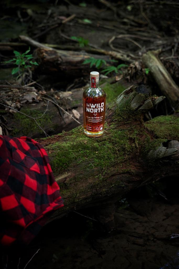 Flasche The Wild North kanadischer Rye Whisky auf einem moosbedeckten Baumstamm im Wald, mit einem karierten rot-schwarzen Hemd im Vordergrund.