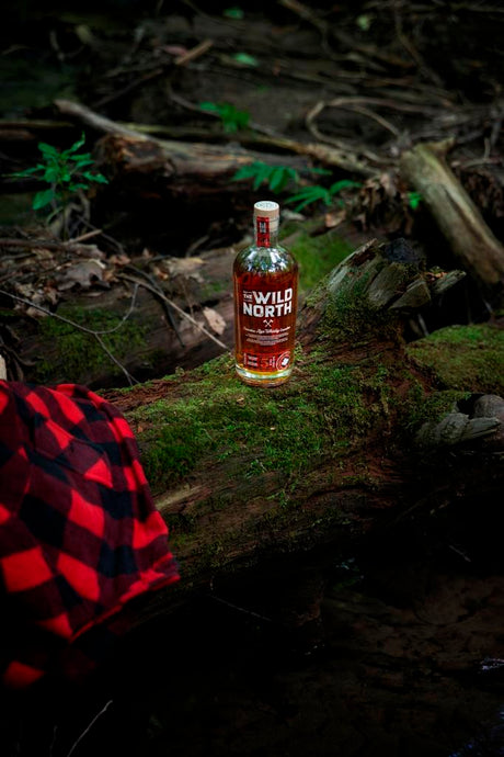 Flasche The Wild North kanadischer Rye Whisky auf einem moosbedeckten Baumstamm im Wald, mit einem karierten rot-schwarzen Hemd im Vordergrund.