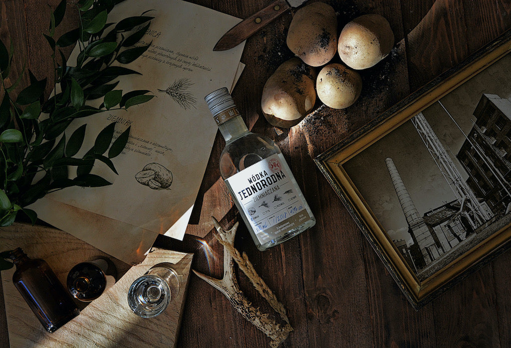 Eine Flasche Wodka Jednorodna Ziemniaczana liegt auf einem Holztisch, umgeben von Kartoffeln, einem Foto im antiken Rahmen, einem Glas und einem Ast.