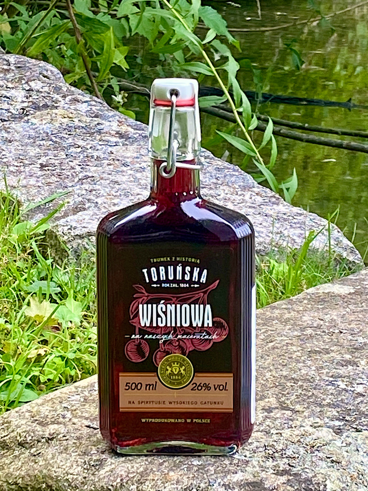 Toruńska Wiśniowa Kirschlikör mit Bügelverschluss auf einem Felsen am See, Flasche mit Kirschmotiv auf dem Etikett.