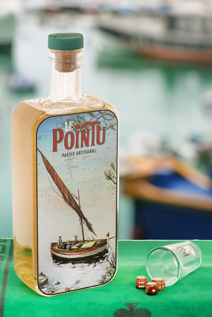 Flasche Pastis Le Pointu auf grünem Tisch mit Shotglas und Würfeln, verschwommener Hafenhintergrund.