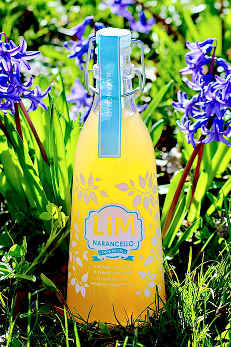 Flasche Lim Narancello Orangenlikör zwischen lila Frühlingsblumen, mit Etikett 'Premium' und Hinweis auf die Herstellung mit mazerierten Orangenschalen. Die Flasche hat eine Bügelverschlusskappe mit einem blauen Band.