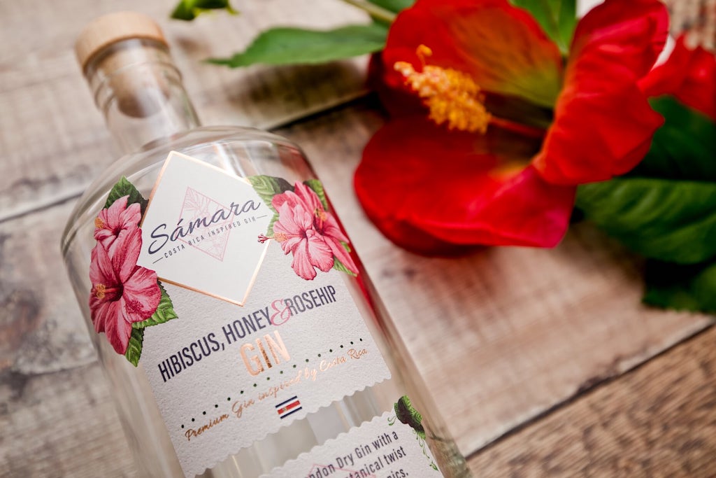 Flasche des Samara Hibiskus, Honig & Hagebutte Gin neben einer roten Blüte, inspiriert von Costa Rica, auf einer Holzoberfläche.