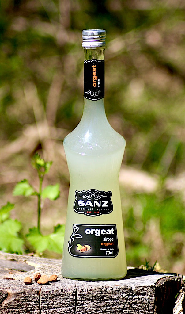 Flasche Sanz Orgeat Sirup, 700 ml, auf einem Baumstumpf mit Mandeln, vor einer natürlichen Waldkulisse.