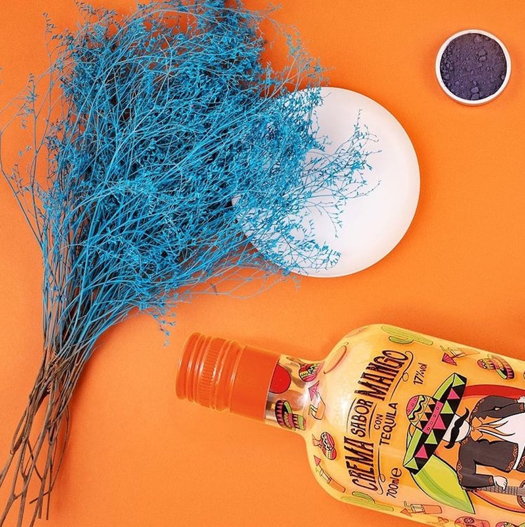 Flasche Mariachi El Mango Tequila-Likör auf orangenem Hintergrund, dekoriert mit blauen getrockneten Blumen und einem weißen Teller.