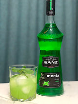 Eine grüne Flasche Minzsirup von Sanz und ein Glas mit grünem Cocktail, Eis und Minzblättern als Garnitur.