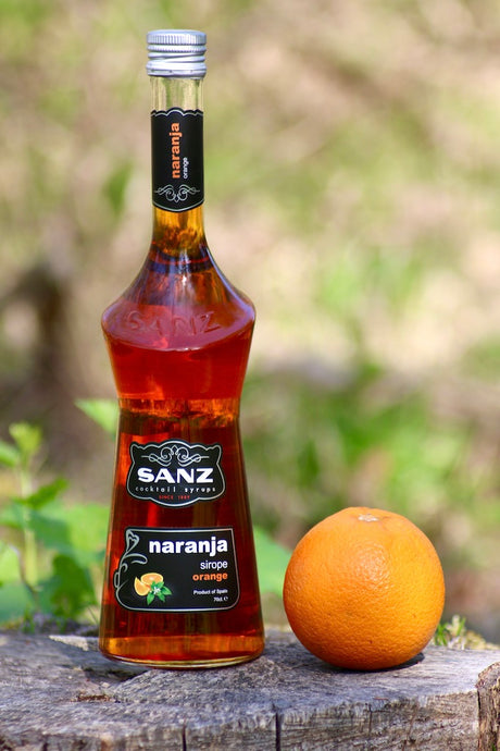 Flasche Sanz Orangensirup mit einer frischen Orange auf einem rustikalen Holzstumpf in natürlicher Umgebung.