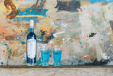 Flasche P'tit Bleu Pastis de Marseille und zwei Gläser mit blauem Getränk vor einem künstlerischen, farblich gesprenkelten Hintergrund.