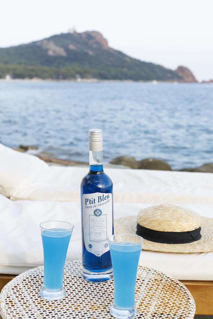 Flasche P'tit Bleu Pastis de Marseille und zwei Gläser mit blauem Getränk auf einem Tisch vor einer Strandkulisse, im Hintergrund ein Hut und das Meer.