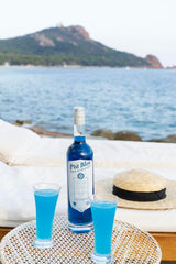 Flasche P'tit Bleu Pastis de Marseille und zwei Gläser mit blauem Getränk auf einem Tisch vor einer Strandkulisse, im Hintergrund ein Hut und das Meer.