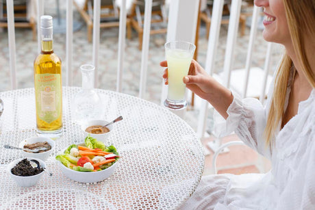 Eine Frau hält ein Glas Pastis neben einer Flasche Le Pastis du Liquoriste, begleitet von Salat und Dips auf einem weißen Tisch.