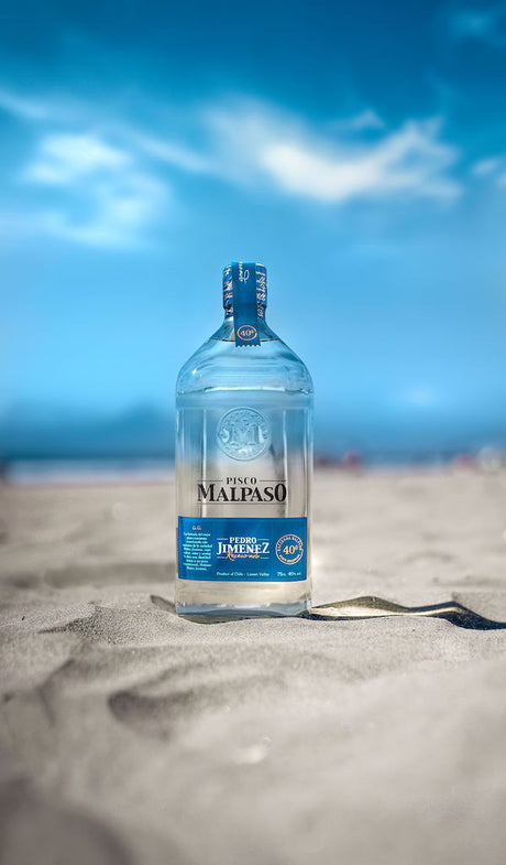 Flasche Pisco MalPaso Pedro Jimenez, 700 ml, 40 % Vol., mit blauem Etikett, stehend auf Sand vor einem blauen Himmel.