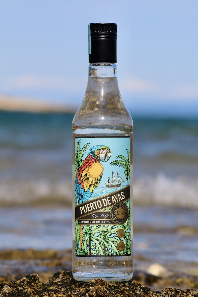 Flasche Puerto de Ayas weißer Rum vor einem unscharfen Meereshintergrund, auf felsigem Untergrund platziert.