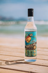 Puerto de Ayas weißer Rum auf einem Holzdeck am Strand mit Meer und blauem Himmel im Hintergrund, tropisches Etikett mit Papagei.