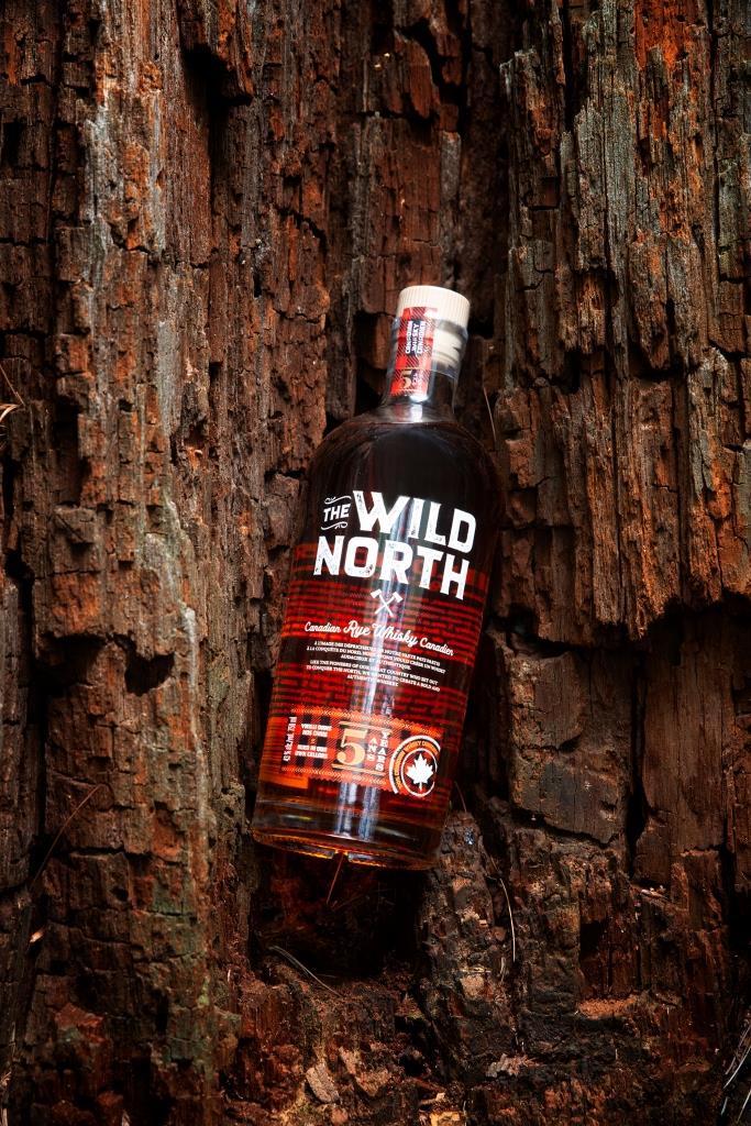 Flasche The Wild North kanadischer Rye Whisky, stehend in einer Vertiefung eines Baumstamms mit rauer, rissiger Rinde. Die braunen Töne des Holzes ergänzen die Farbe des Whiskys.