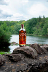 Flasche The Wild North kanadischer Rye Whisky, auf einem Felsen platziert, im Hintergrund ein Fluss und bewaldete Ufer. Der Himmel ist leicht bewölkt, und die Naturumgebung schafft eine ruhige Atmosphäre