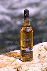 Eine Flasche Crac's Blended Scotch Whisky, stehend auf einem Felsen vor einem unscharfen Flusshintergrund. Die Flasche zeigt golden schimmernden Whisky und ein Etikett mit einem schottischen Schloss.