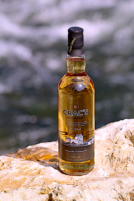 Eine Flasche Crac's Blended Scotch Whisky, stehend auf einem Felsen vor einem unscharfen Flusshintergrund. Die Flasche zeigt golden schimmernden Whisky und ein Etikett mit einem schottischen Schloss.