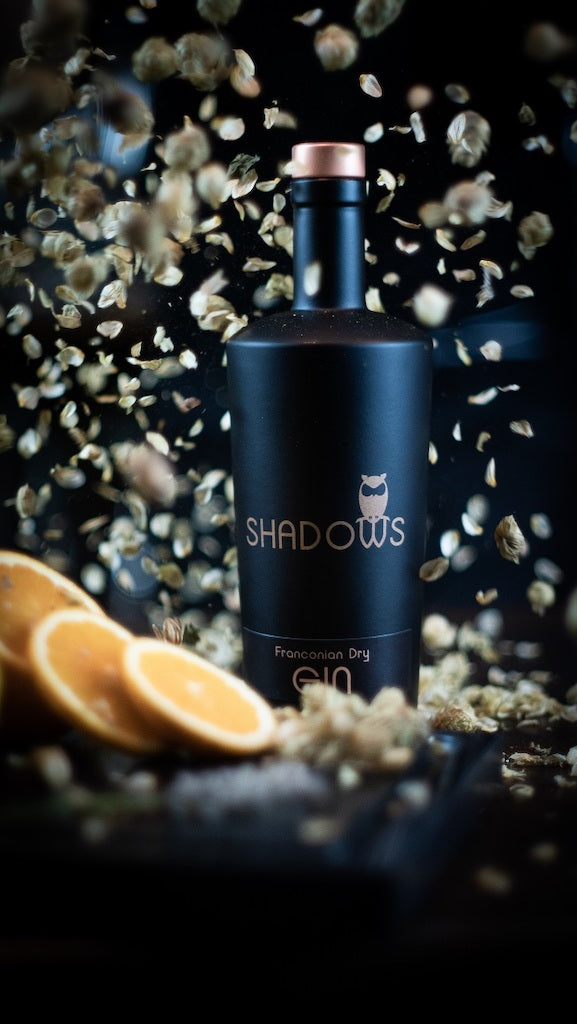 Shadows Franconian Dry Gin Flasche mit umherfliegenden Hopfenblüten und Orangenscheiben, stilvoll in Szene gesetzt.