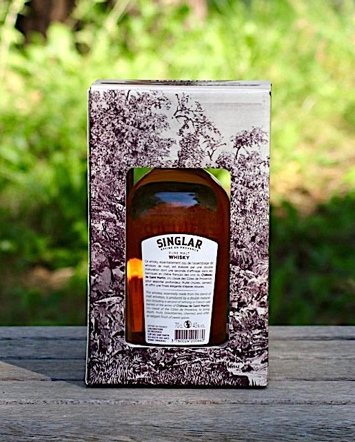 Rückseite der Geschenkbox des Singlar Pure Malt Whisky, mit Blick auf das Etikett der Flasche. Die Verpackung zeigt eine detaillierte Illustration, präsentiert vor einem unscharfen grünen Hintergrund.