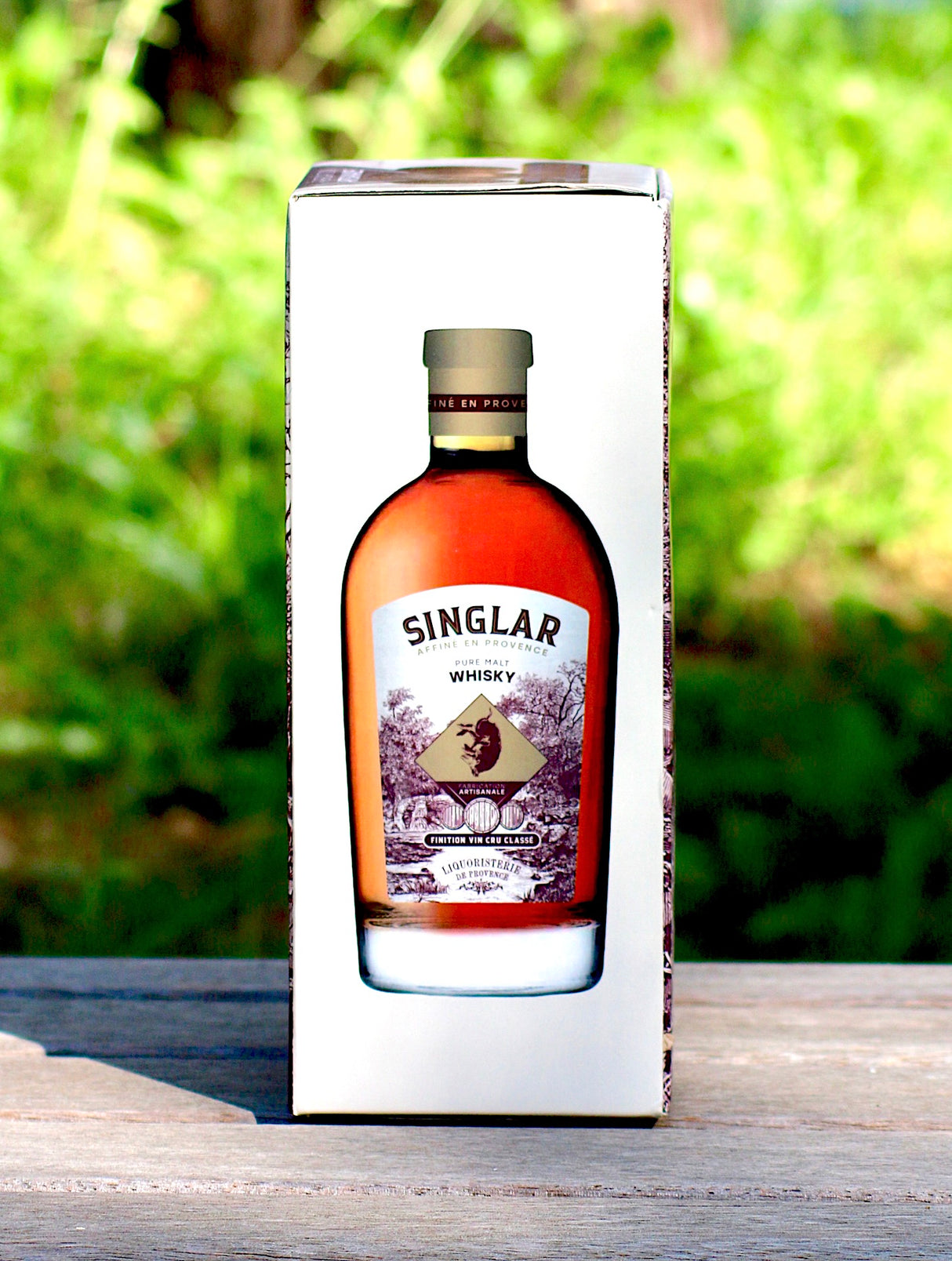 Seitenansicht der Geschenkbox des Singlar Pure Malt Whisky, mit einer Abbildung der Flasche auf der Seite der Verpackung. Der Whisky wurde in der Provence veredelt, präsentiert vor einem unscharfen, grünen Hintergrund.