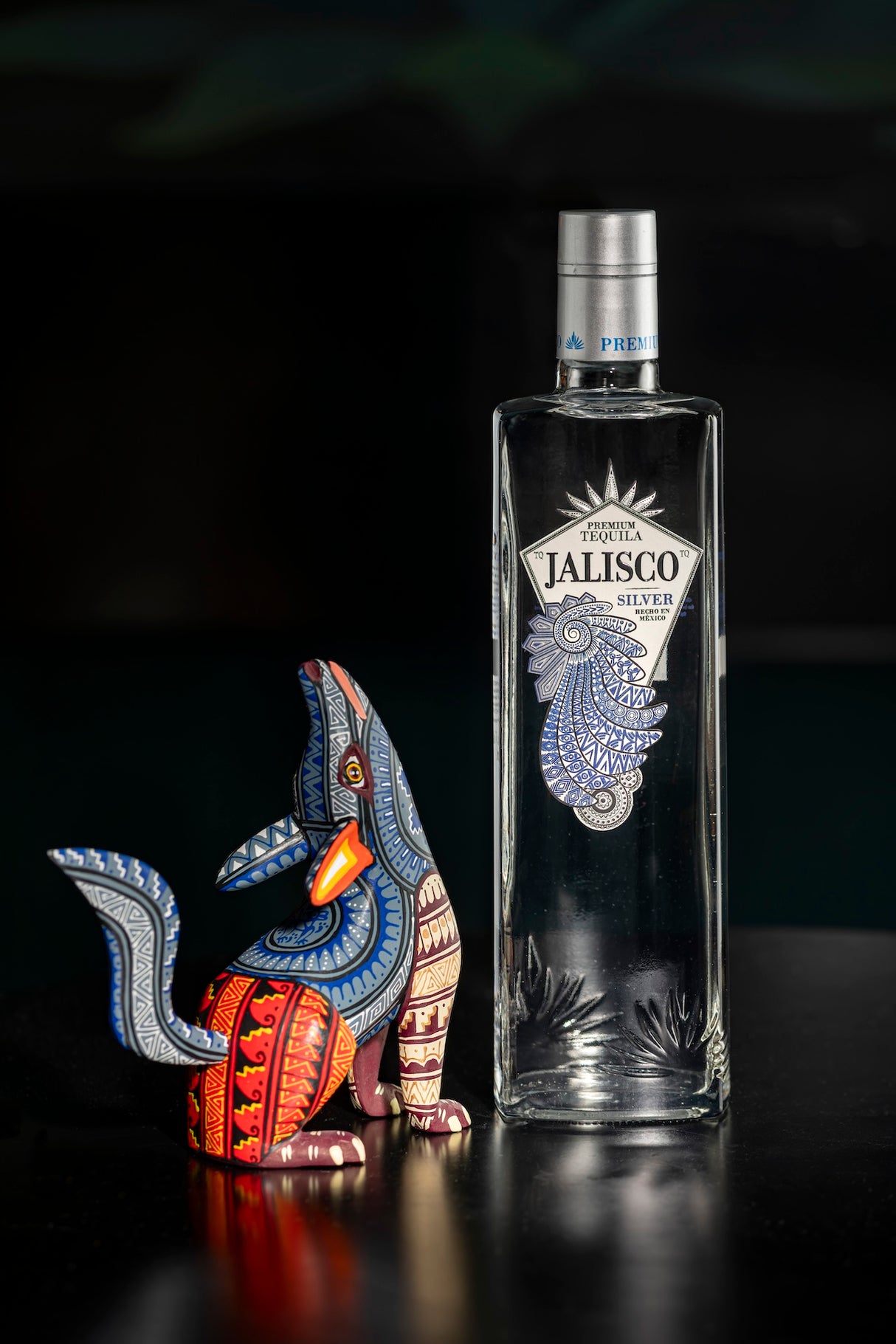 Tequila-Jalisco-Silver-Flasche neben einer bunt bemalten Alebrije-Holzfigur in Tierform auf dunklem Hintergrund.