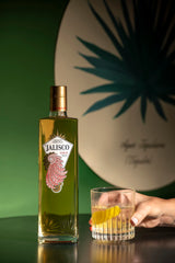 Tequila-Jalisco-Gold-Flasche neben einem Glas mit gelber Zeste, gehalten von einer Hand vor grünem Agavenmotiv.