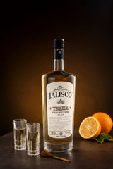 Flasche Tequila Jalisco Reposado mit zwei gefüllten Shotgläsern, Zimtstangen und einer halbierten Orange im Hintergrund.