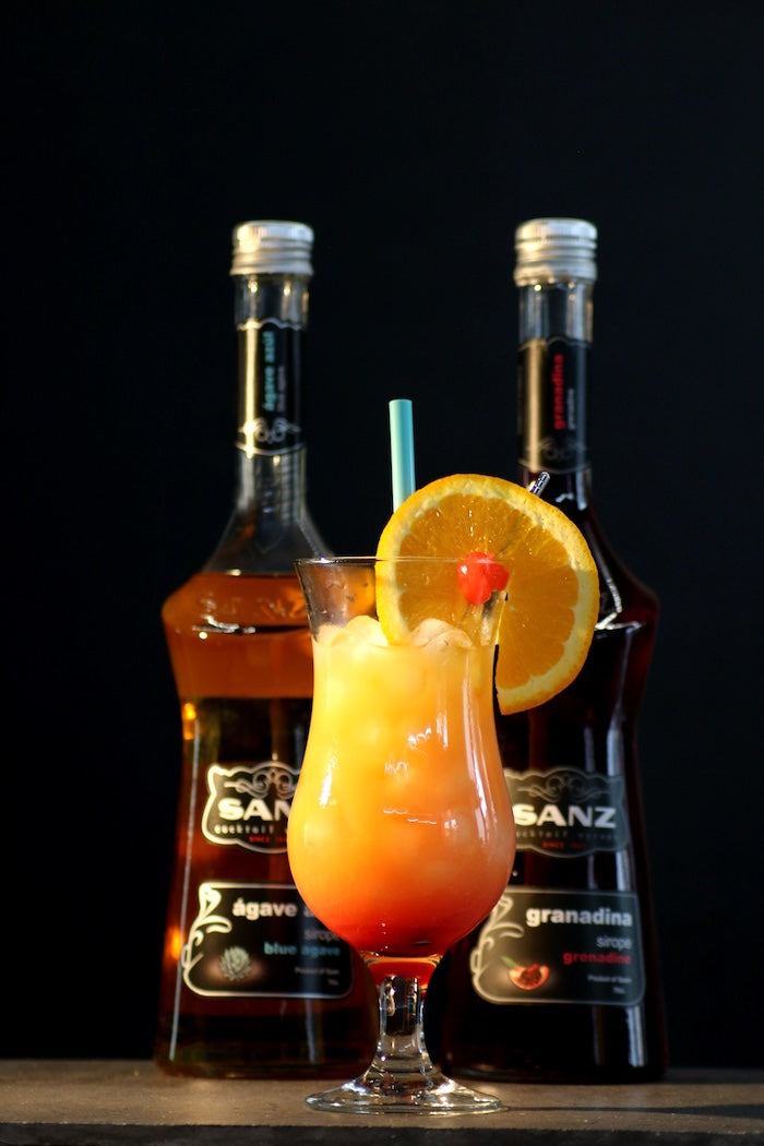 Alkoholfreier Tequila Sunrise mit Sanz Grenadine und Agavensirup, dekoriert mit Orangenscheibe und Kirsche, Flaschen im Hintergrund.