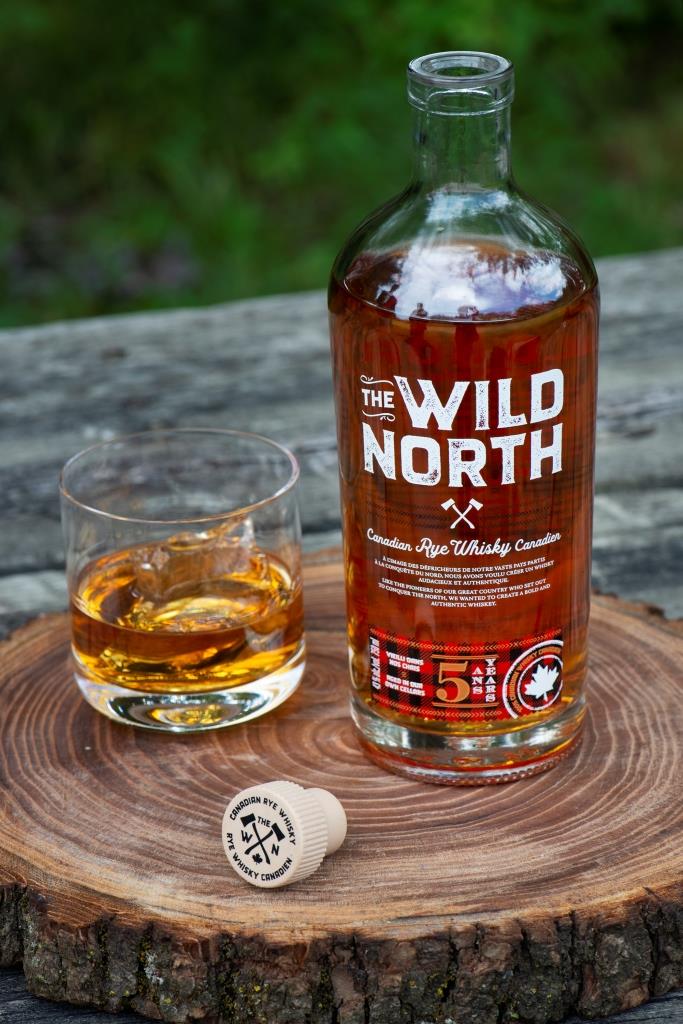 Flasche The Wild North kanadischer Rye Whisky auf einem Holzbrett, daneben ein Glas Whisky mit Eiswürfeln und ein Korken. Im Hintergrund ist eine natürliche grüne Umgebung zu sehen.