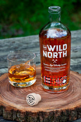 Flasche The Wild North kanadischer Rye Whisky auf einem Holzbrett, daneben ein Glas Whisky mit Eiswürfeln und ein Korken. Im Hintergrund ist eine natürliche grüne Umgebung zu sehen.