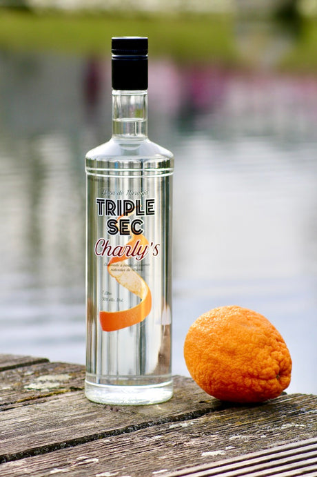 Flasche Triple Sec Charly’s auf Holzsteg vor Wasser, daneben eine ganze Orange.